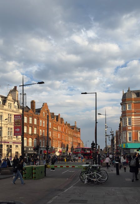 The image depicts a busy urban street scene in Kentish Town, featuring a historic brick building with large arched windows and a small tower at the top, set against a clear blue sky. In the foreground