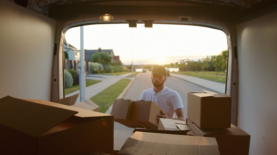 A man wearing a white polo shirt and glasses is standing outside a white moving van with its rear doors open during a home relocation. He is holding a cardboard box and appears to be in the process of