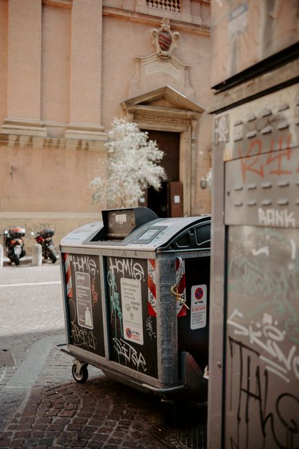 A large green wheeled waste bin with the number 53 and the label 'JUN BESAR' is positioned on the pavement in front of a storage area containing shelves with various household items, including boxes,