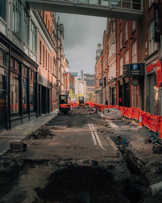 A city street undergoing roadwork with the pavement dug up, exposing uneven dirt and construction debris. Red plastic barriers line the edges of the excavation area, preventing access, while a small excavator is visible in the distance. On the left side, there are shops with large glass windows, and on the right side, multi-storey brick buildings with signage, including a 'P' parking sign. The scene is viewed from beneath a bridge or overpass, with the underside visible at the top of the image. The lighting is natural, with overcast sky casting diffuse light across the scene. This setting illustrates the logistics and challenges of home relocation or furniture transport in urban environments, where road access may be temporarily restricted during partial road closures managed by [COMPANY_NAME] for residential or commercial moves in the area related to the Kentish Town Road removals guide for narrow access.
