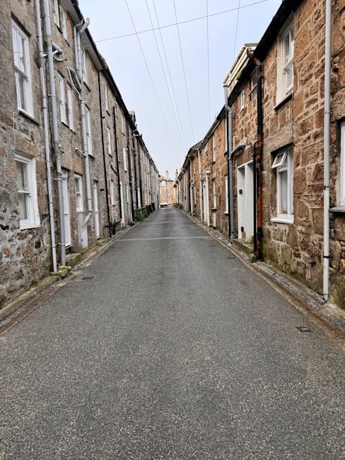 A city street undergoing roadwork with the pavement dug up, exposing uneven dirt and construction debris. Red plastic barriers line the edges of the excavation area, preventing access, while a small excavator is visible in the distance. On the left side, there are shops with large glass windows, and on the right side, multi-storey brick buildings with signage, including a 'P' parking sign. The scene is viewed from beneath a bridge or overpass, with the underside visible at the top of the image. The lighting is natural, with overcast sky casting diffuse light across the scene. This setting illustrates the logistics and challenges of home relocation or furniture transport in urban environments, where road access may be temporarily restricted during partial road closures managed by [COMPANY_NAME] for residential or commercial moves in the area related to the Kentish Town Road removals guide for narrow access.