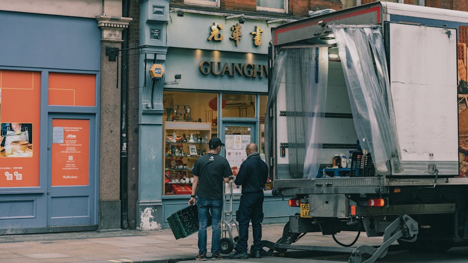 A white moving van with its rear door open, parked on a busy town street in Kentish Town. Inside the van, partially visible packing materials and boxes are ready for loading or unloading. The vehicle