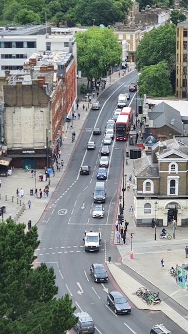 A street scene in Kentish Town showing a historic brick building with large arched windowpanes on the left, and a blue bridge with a yellow 'Camden Lock' sign spanning the road ahead. The sidewalk is